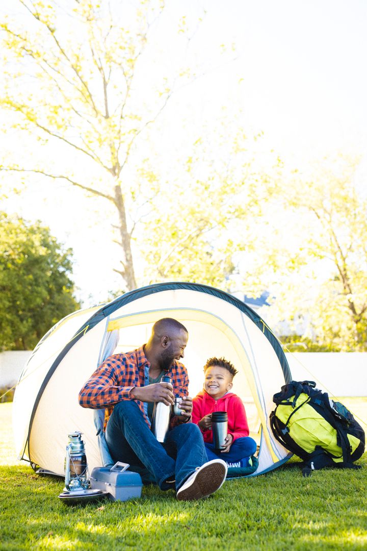 Happy african american father and son sitting in tent and drinking from thermos in their backyard - Free Images, Stock Photos and Pictures on Pikwizard.com