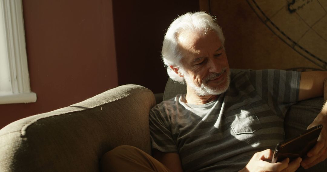 Elderly Man Relaxing on Sofa Using Smartphone in Sunlit Room - Free Images, Stock Photos and Pictures on Pikwizard.com