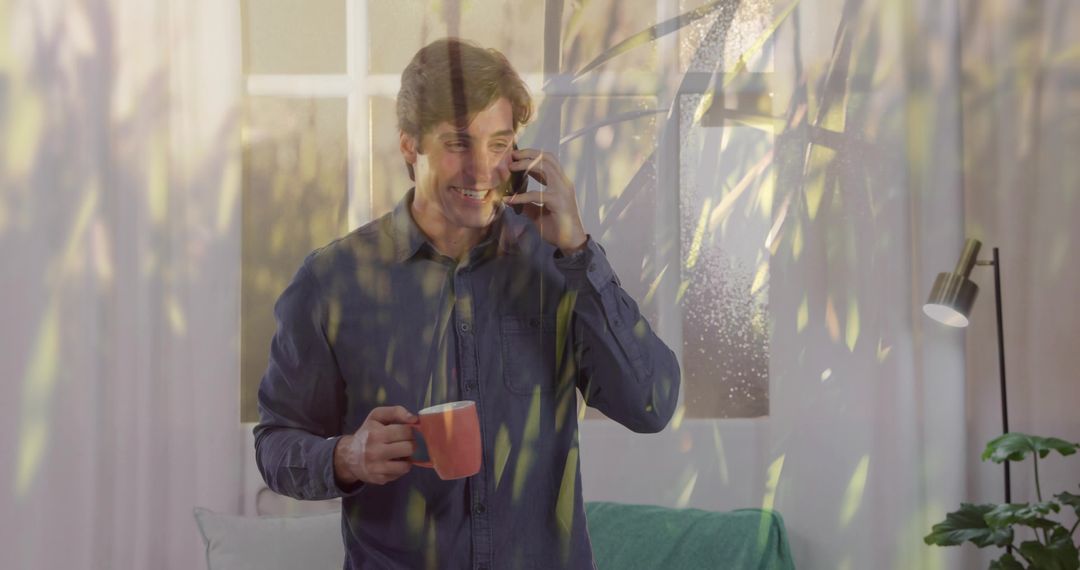 Smiling man talking on smartphone while holding orange mug in sunlit living room