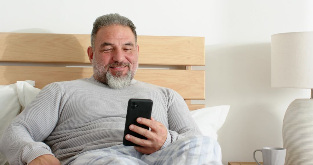 Senior man smiling while using smartphone in bed, relaxing cozy morning lifestyle