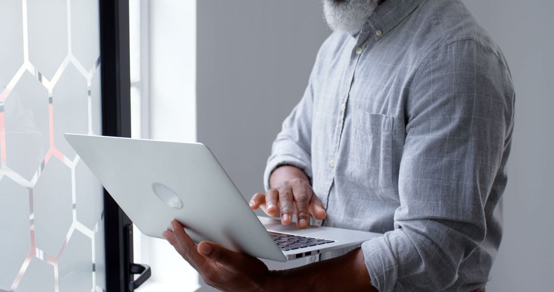 Older Man Using Laptop by Window in Bright Office Setting - Free Images, Stock Photos and Pictures on Pikwizard.com