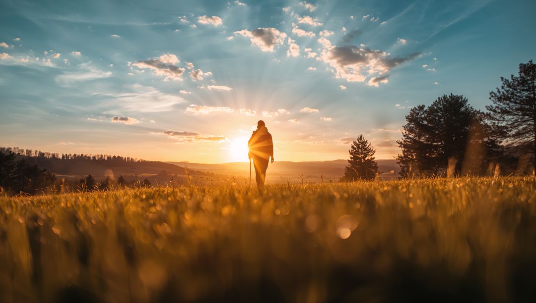 Silhouetted hiker walking into golden sunrise across dew-covered meadow with sunbeams