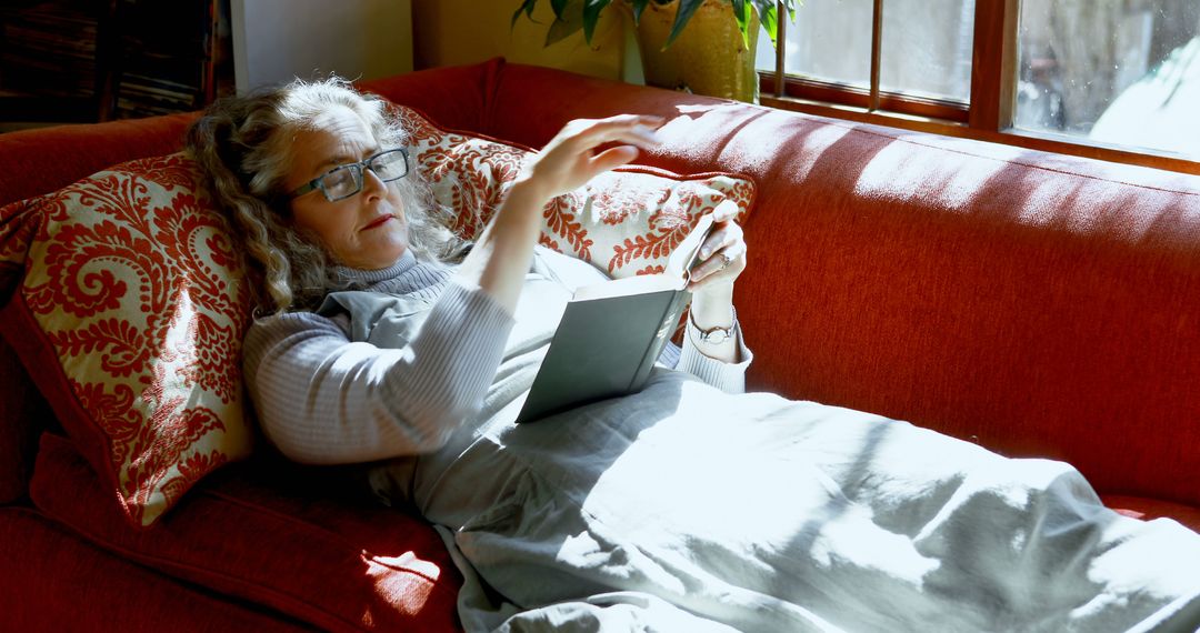 Senior Woman Reading Book on Cozy Red Sofa in Sunlit Living Room - Free Images, Stock Photos and Pictures on Pikwizard.com