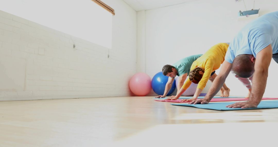 Group practicing downward dog yoga in bright studio with stability balls and sunlight