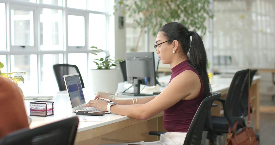 Indian woman working on laptop at open-plan office desk, modern bright coworking workspace