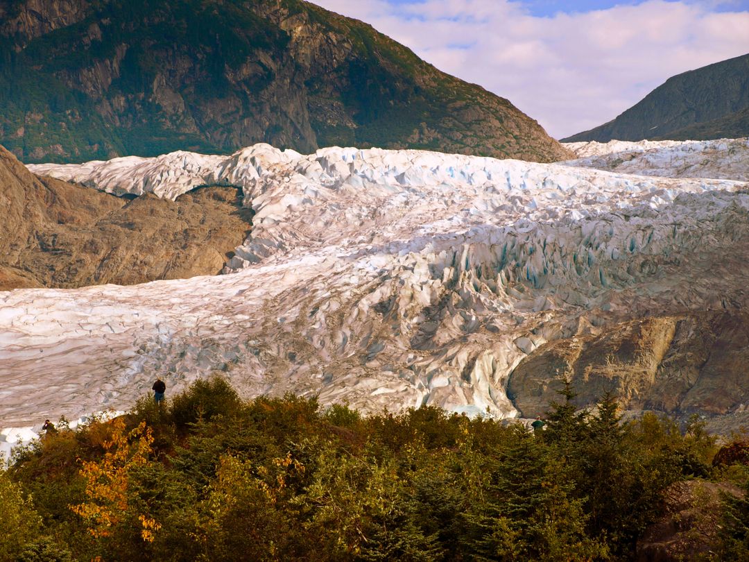 Hiker watching vast glacier flowing through rugged mountain valley with autumn foreground
