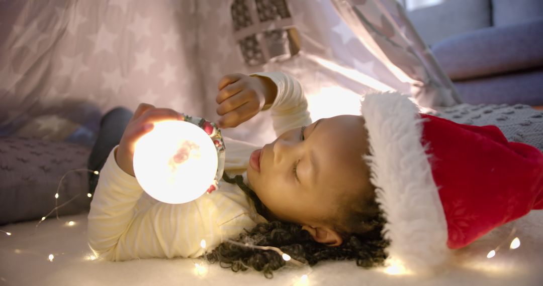 Young girl in Santa hat lying under cozy blanket playing with lights - Free Images, Stock Photos and Pictures on Pikwizard.com