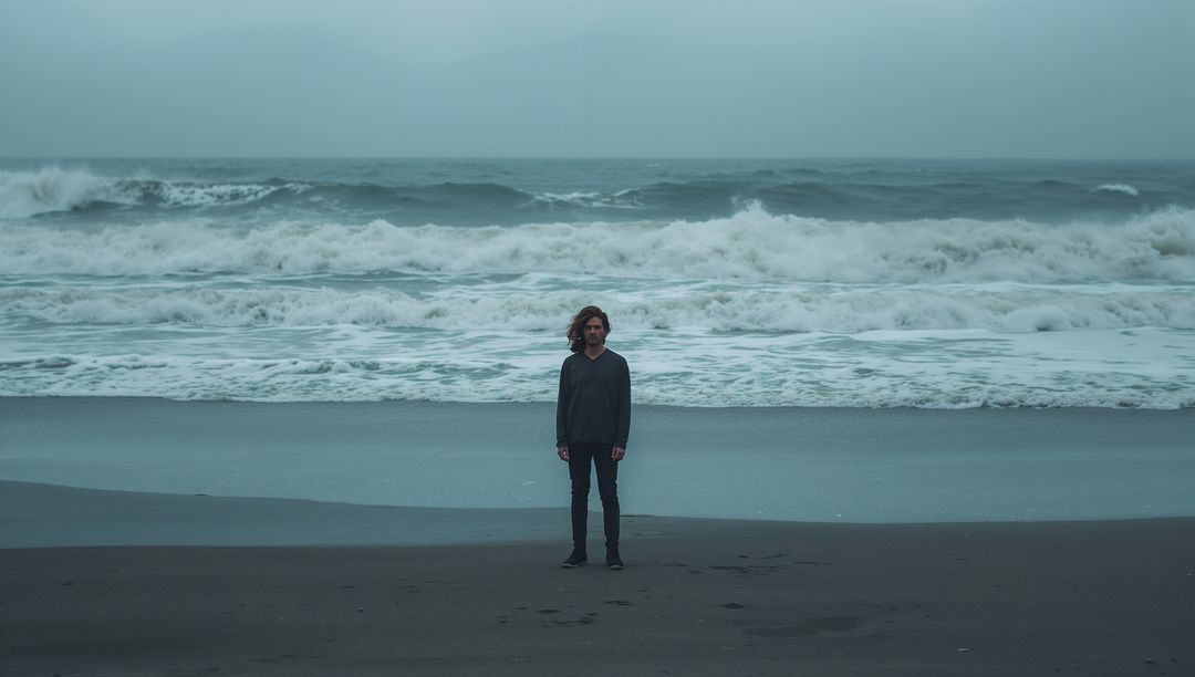 Solitary man standing on moody dark beach facing camera with crashing waves and overcast sky