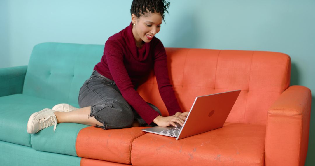 Smiling woman typing on laptop while lounging on teal and orange sofa for remote work