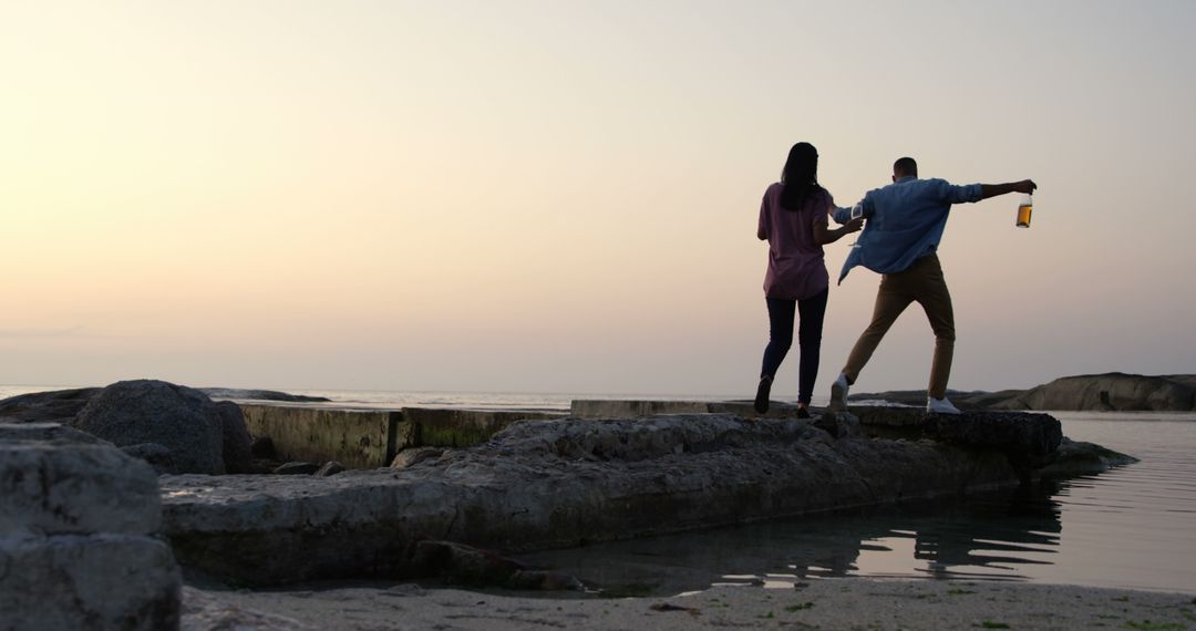 Biracial couple balancing on rocks by sea, holding hands - Free Images, Stock Photos and Pictures on Pikwizard.com
