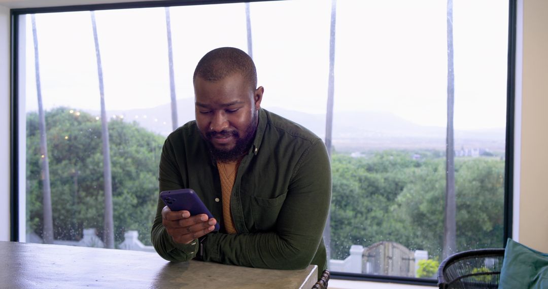 African American man leaning on kitchen counter using smartphone by large window wearing earbud