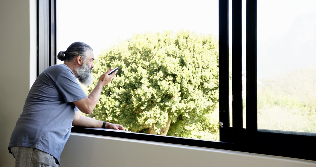 Senior Man Using Smartphone Near Large Open Window