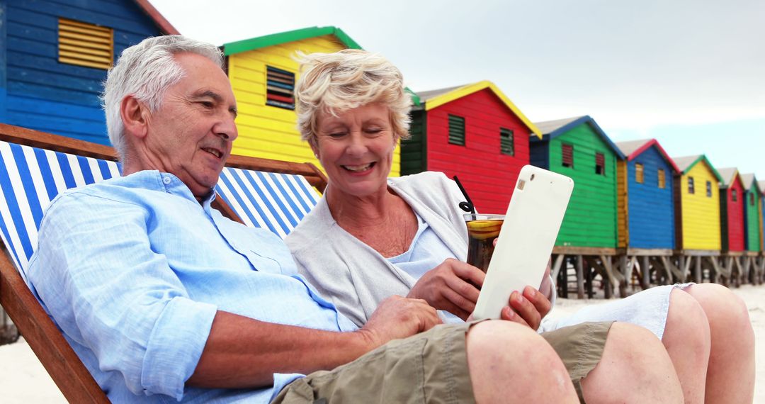 Senior Couple Relaxing on Beach with Tablet in Front of Colorful Beach Huts - Free Images, Stock Photos and Pictures on Pikwizard.com