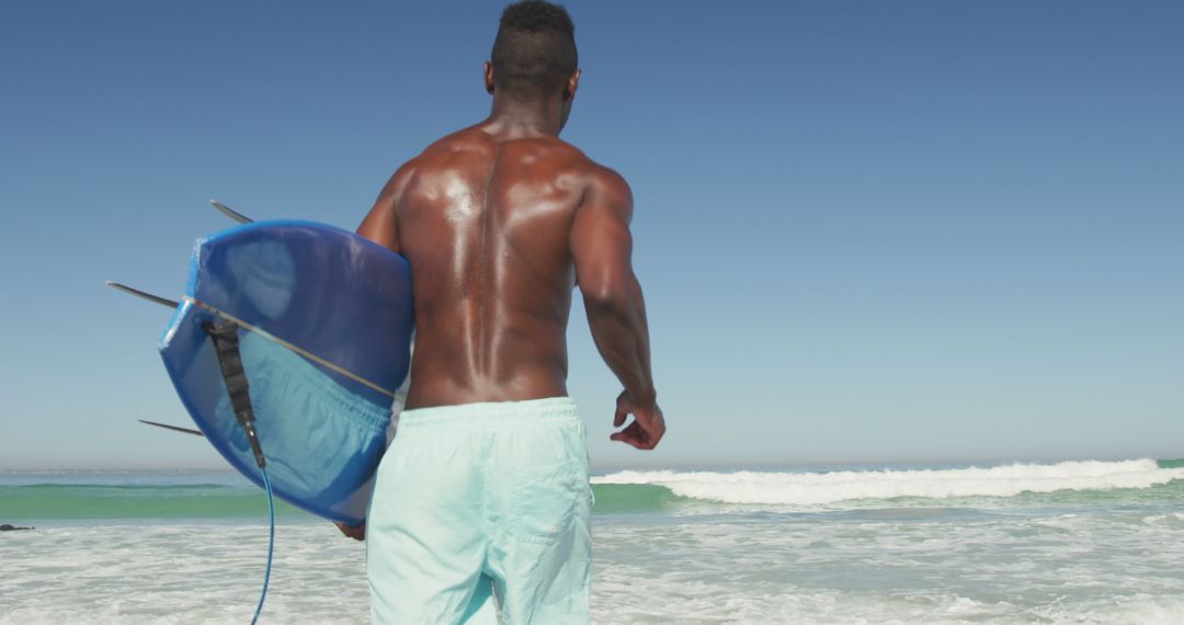 Rear view of african american man carrying surfboard running on sunny beach into the sea - Free Images, Stock Photos and Pictures on Pikwizard.com