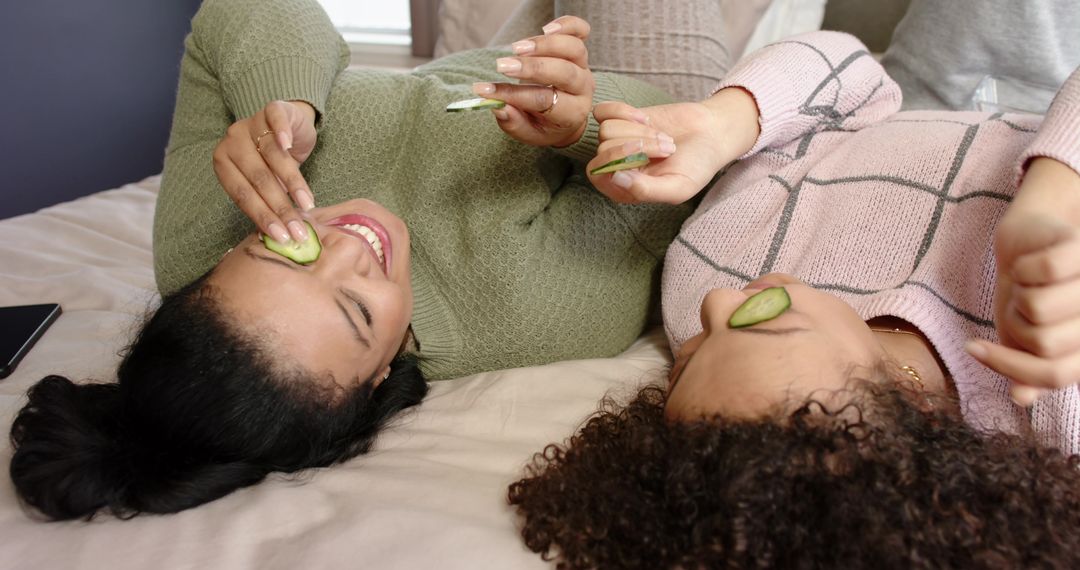 Friends Lying on Bed with Cucumber Eye Slices Enjoying Home Spa, Cozy Sweaters, Laughing