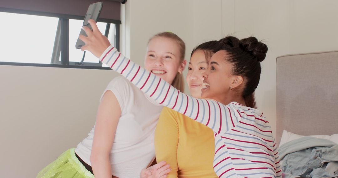 Diverse friends smiling while taking group selfie on bed bathed in natural light