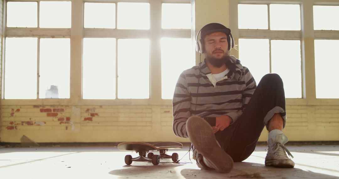 Young man relaxing with skateboard in sunlit loft wearing headphones - Free Images, Stock Photos and Pictures on Pikwizard.com