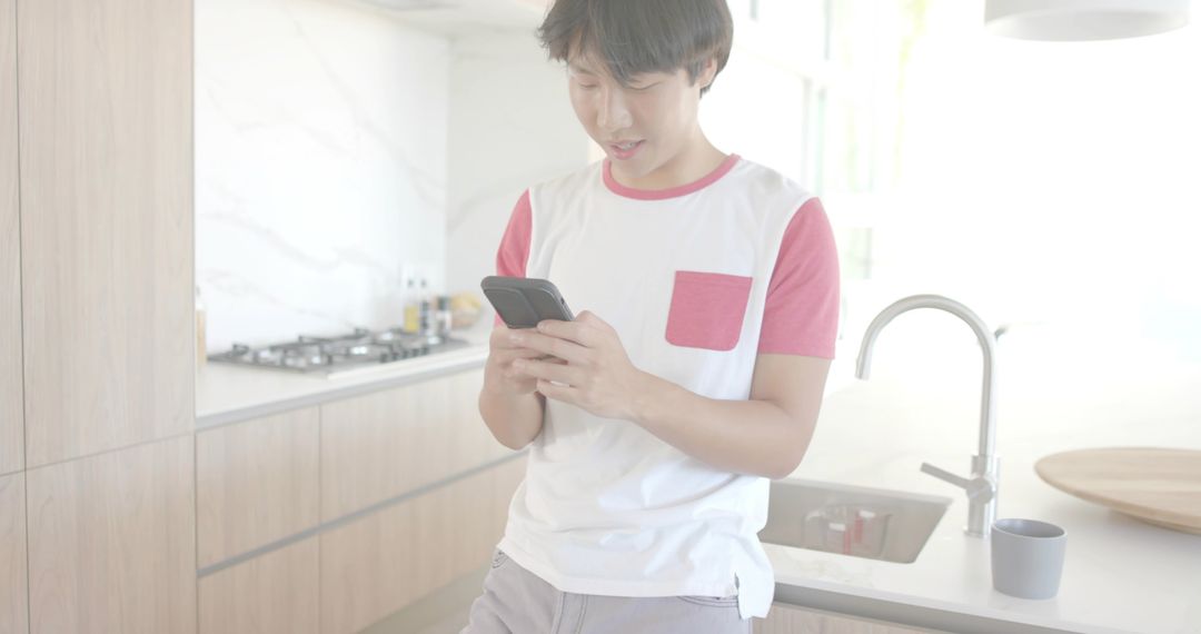 Leaning on counter checking smartphone in bright minimalist kitchen, young man lifestyle