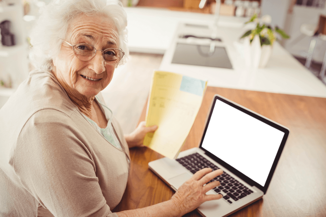 Elderly Woman Smiling, Using Laptop and Holding Document at Home Table