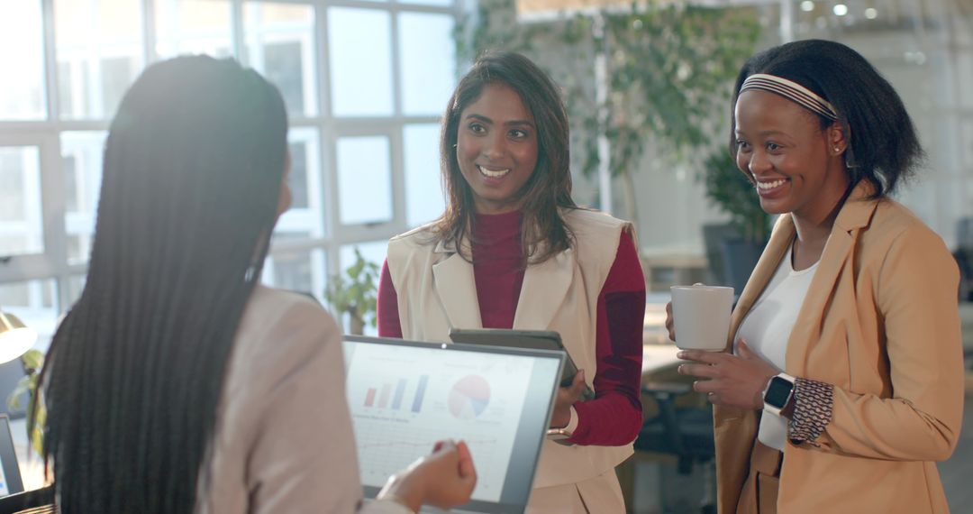 Diverse female team collaborating around tablet and laptop with analytics in bright office
