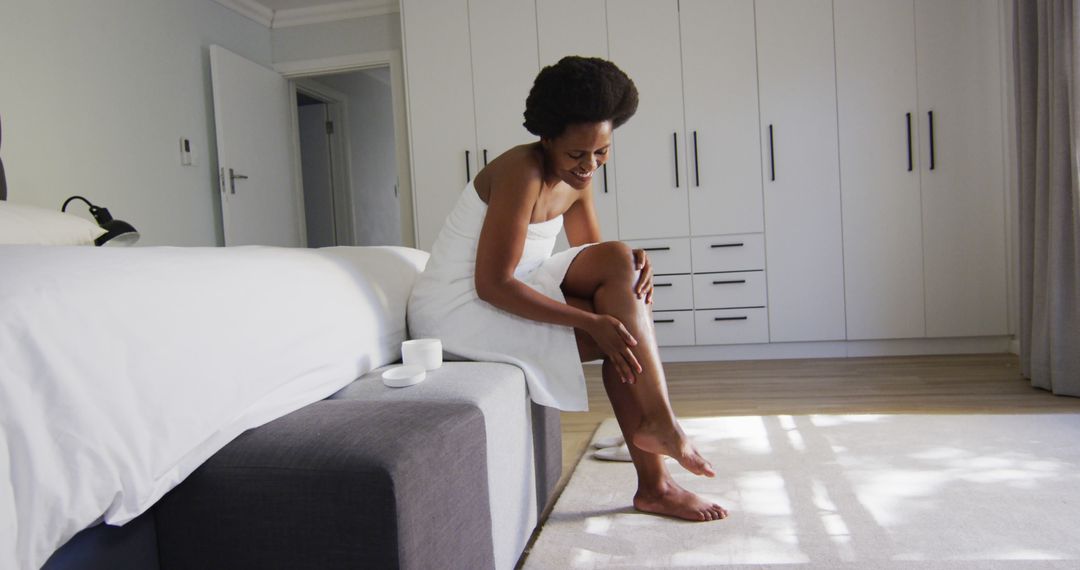 Happy african american woman apply cream on legs in bedroom - Free Images, Stock Photos and Pictures on Pikwizard.com