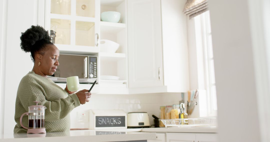 Woman Checking Smartphone while Holding Mug in White Kitchen - Free Images, Stock Photos and Pictures on Pikwizard.com