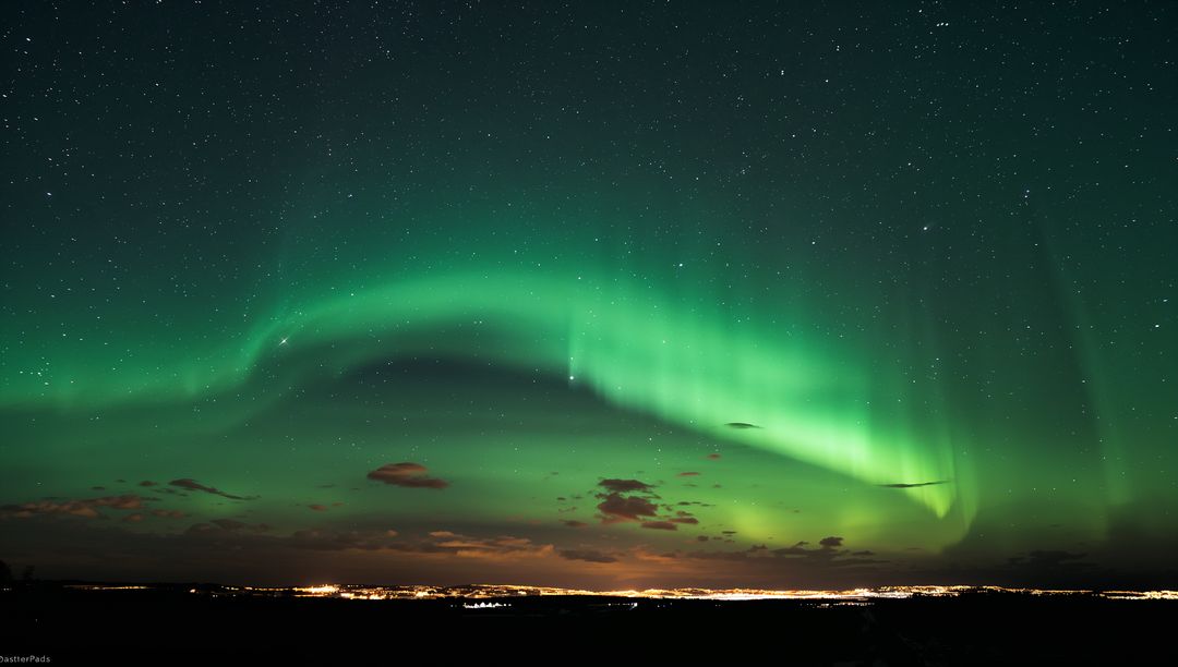 Emerald aurora arching across starry night above distant town lights and dark silhouette