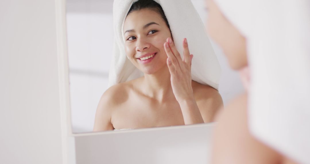Image of portrait of smiling biracial woman with towel on hair applying serum in bathroom - Free Images, Stock Photos and Pictures on Pikwizard.com
