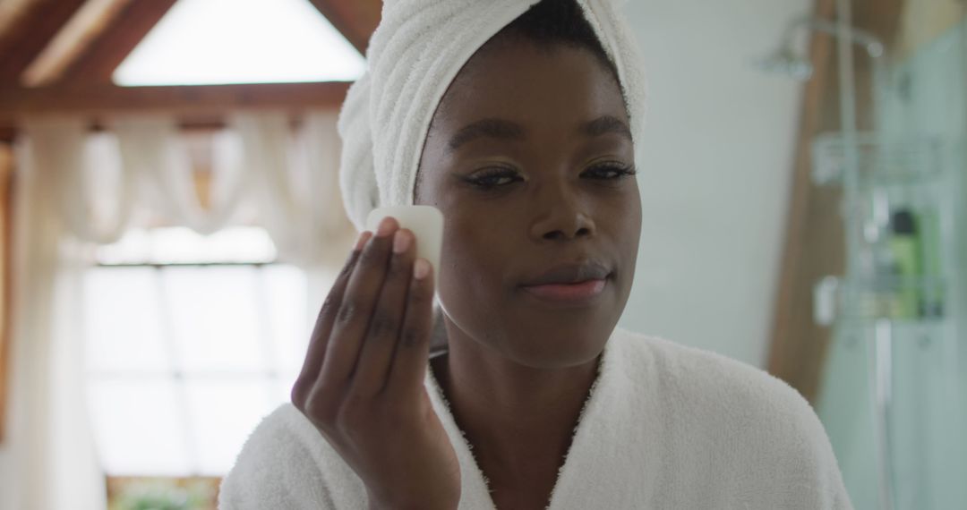 Woman wearing bathrobe applying skincare product with cotton pad in bathroom - Free Images, Stock Photos and Pictures on Pikwizard.com