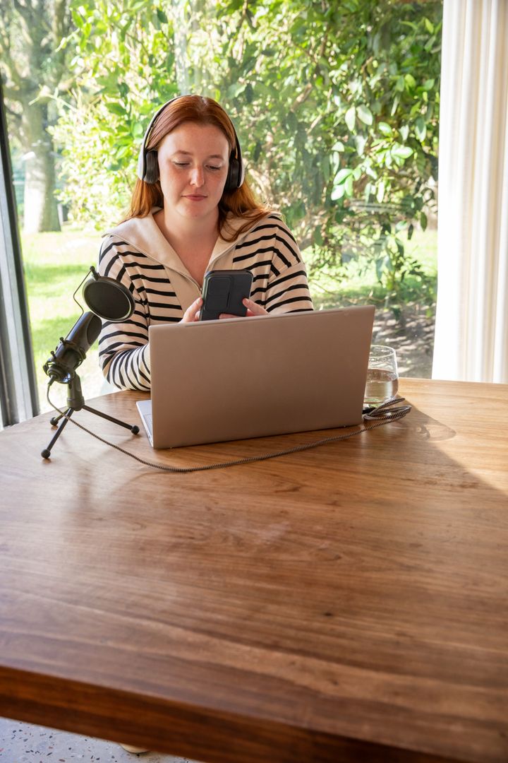 Woman Using Smartphone in Home Office with Laptop and Microphone on Table