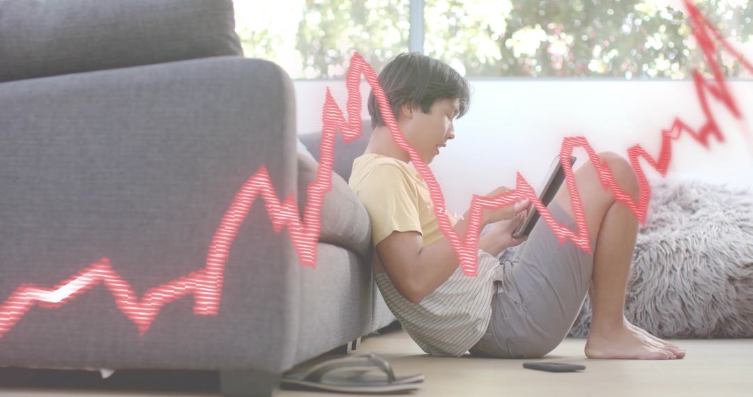 Asian man using tablet on floor beside sofa with red stock chart overlay in living room