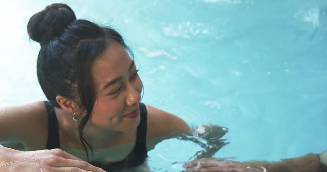Asian woman smiling in turquoise pool leaning on edge in black swimsuit with hoop earring