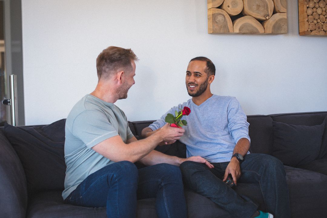 Diverse gay male couple sitting on sofa one giving red rose to the other - Free Images, Stock Photos and Pictures on Pikwizard.com