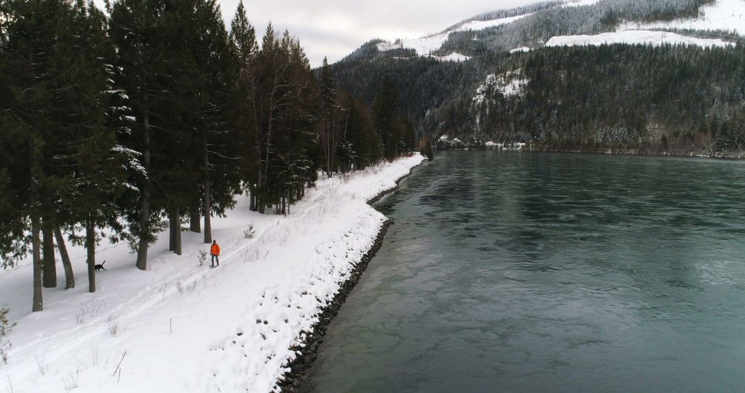 Lone Traveler Walking along Snow-covered Path beside Tranquil River in Winter Landscape - Free Images, Stock Photos and Pictures on Pikwizard.com