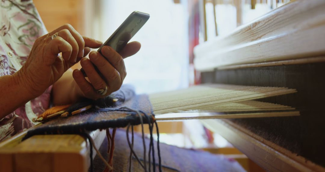 Senior Woman Weaving on Handloom and Using Smartphone in Workshop