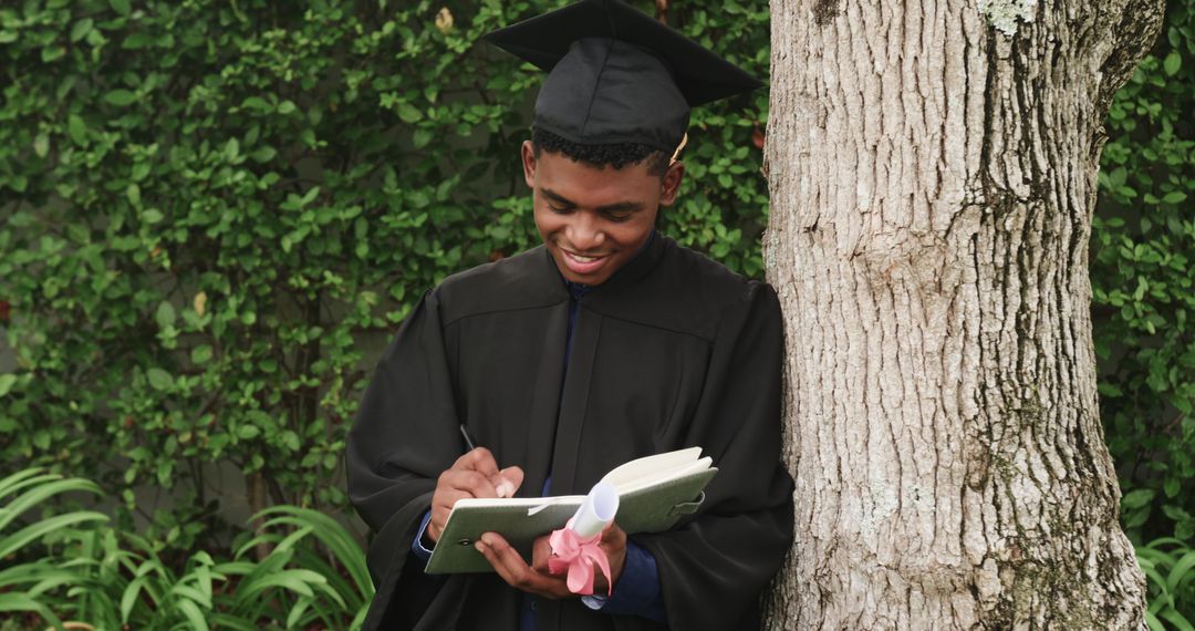 African American Graduate Writing Notebook While Leaning on Tree and Holding Diploma