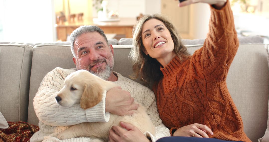 Smiling couple taking cozy selfie on sofa with golden retriever puppy in knit sweaters