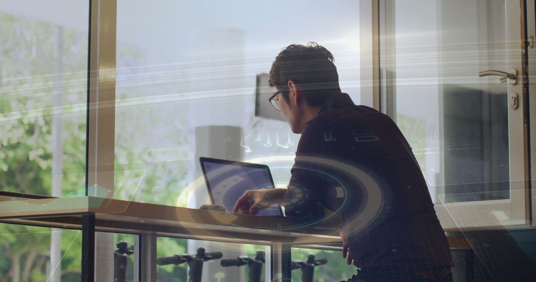 Focused Young Man Typing on Laptop Near Window with Digital Overlay