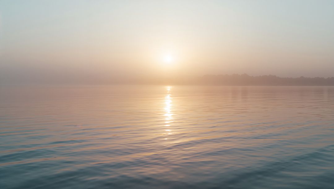 Glowing sun creating vertical reflection on calm lake at dawn with misty treeline