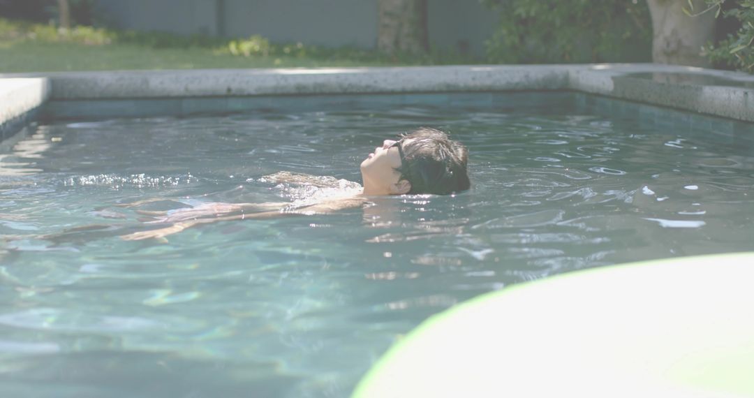 Sunlit backyard pool floating Asian man wearing sunglasses relaxing on back near inflatable