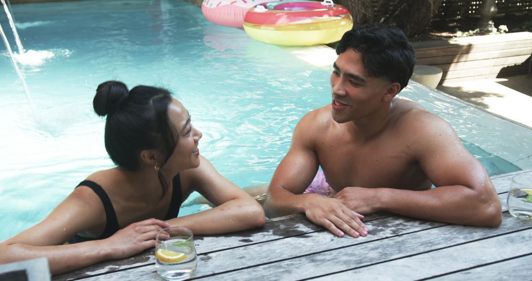 Young couple leaning and chatting at backyard pool on wooden deck with lemon drinks