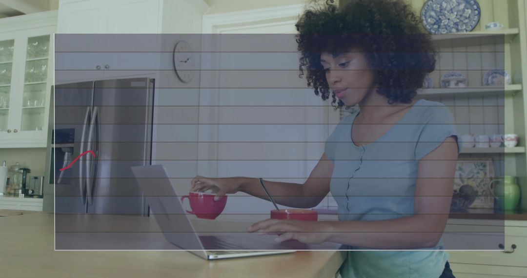 Woman working on laptop at kitchen island stirring red mug multitasking in modern kitchen