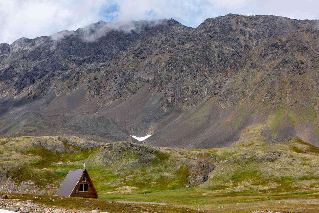 Triangle-shaped cabin nesting beneath rocky mountain in mossy alpine tundra panorama