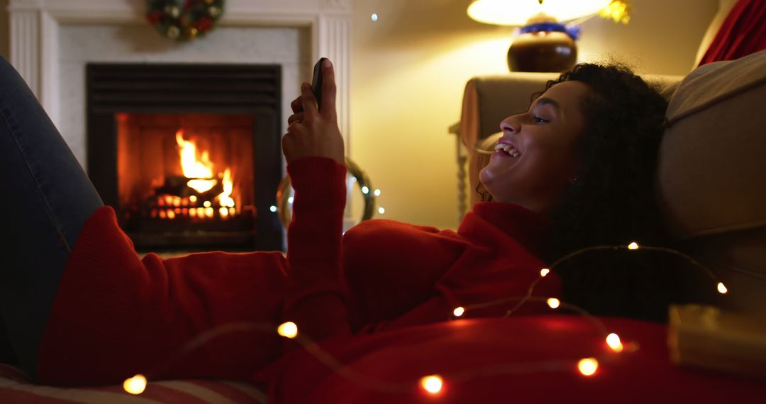 Woman smiling and using smartphone by fireplace during holiday season - Free Images, Stock Photos and Pictures on Pikwizard.com