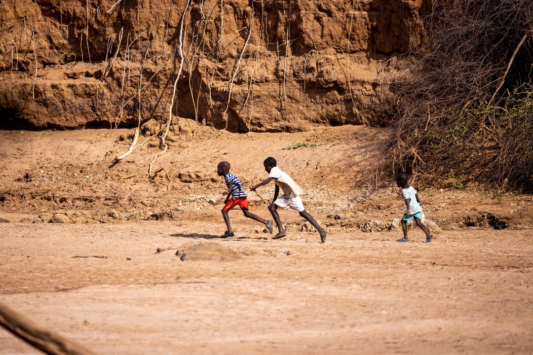 Three boys running across dry riverbed near eroded clay cliffs, playing together outdoors
