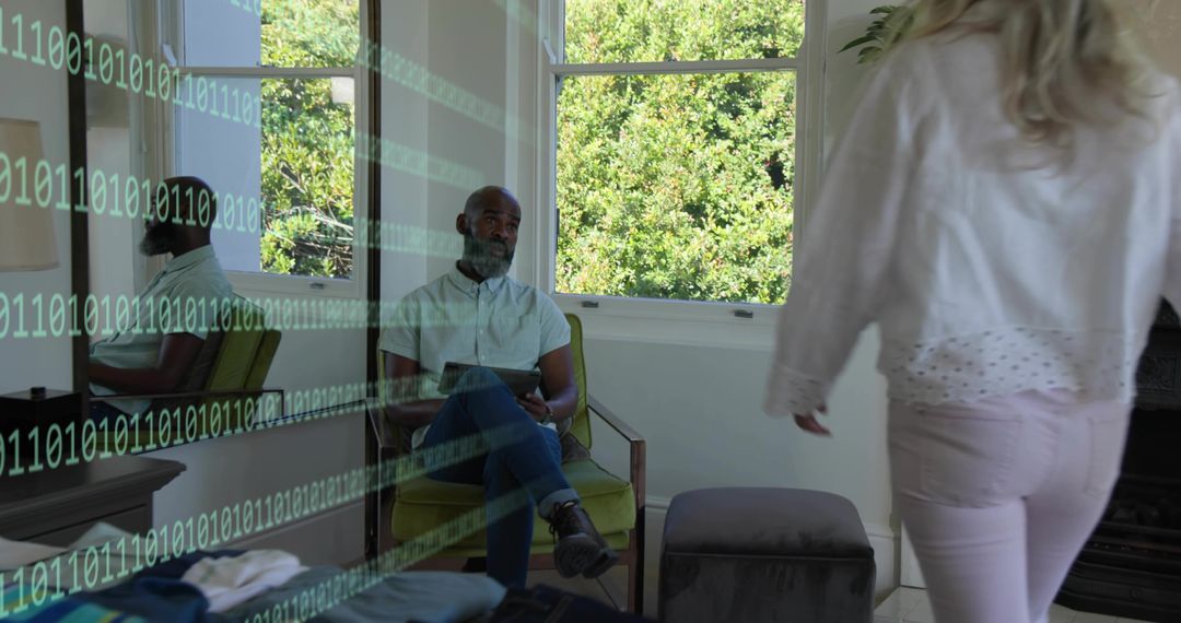 Man Sitting with Tablet in Sunlit Living Room While Woman Walks Past