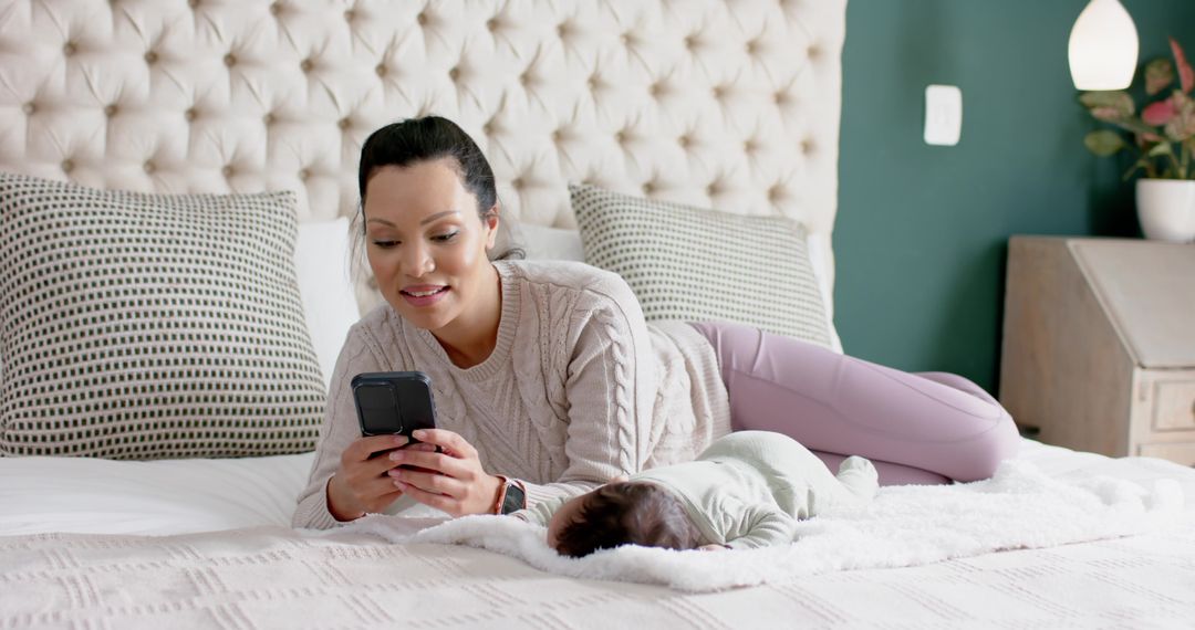 Mother Relaxing with Smartphone Beside Resting Infant in Bedroom