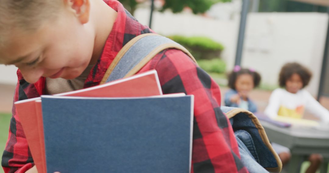 Young Boy Carrying Books Outdoors While Classmates Study In Background - Free Images, Stock Photos and Pictures on Pikwizard.com