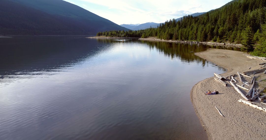 Solo Traveler Relaxing on Secluded Lakeside Beach with Forested Mountains - Free Images, Stock Photos and Pictures on Pikwizard.com