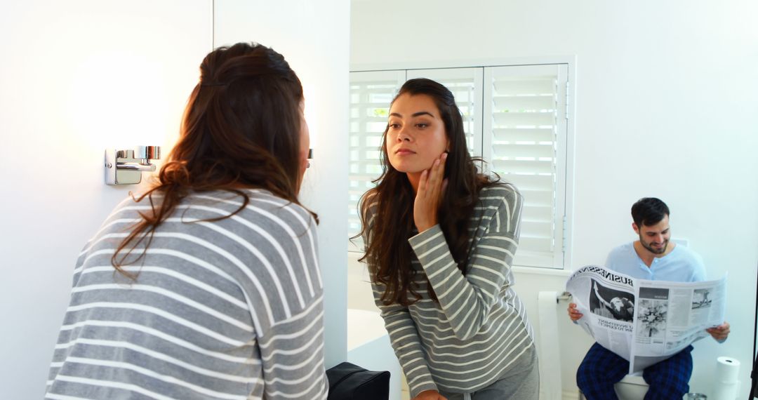 Young Woman Examining Skin in Bathroom Mirror as Man Reads Newspaper - Free Images, Stock Photos and Pictures on Pikwizard.com